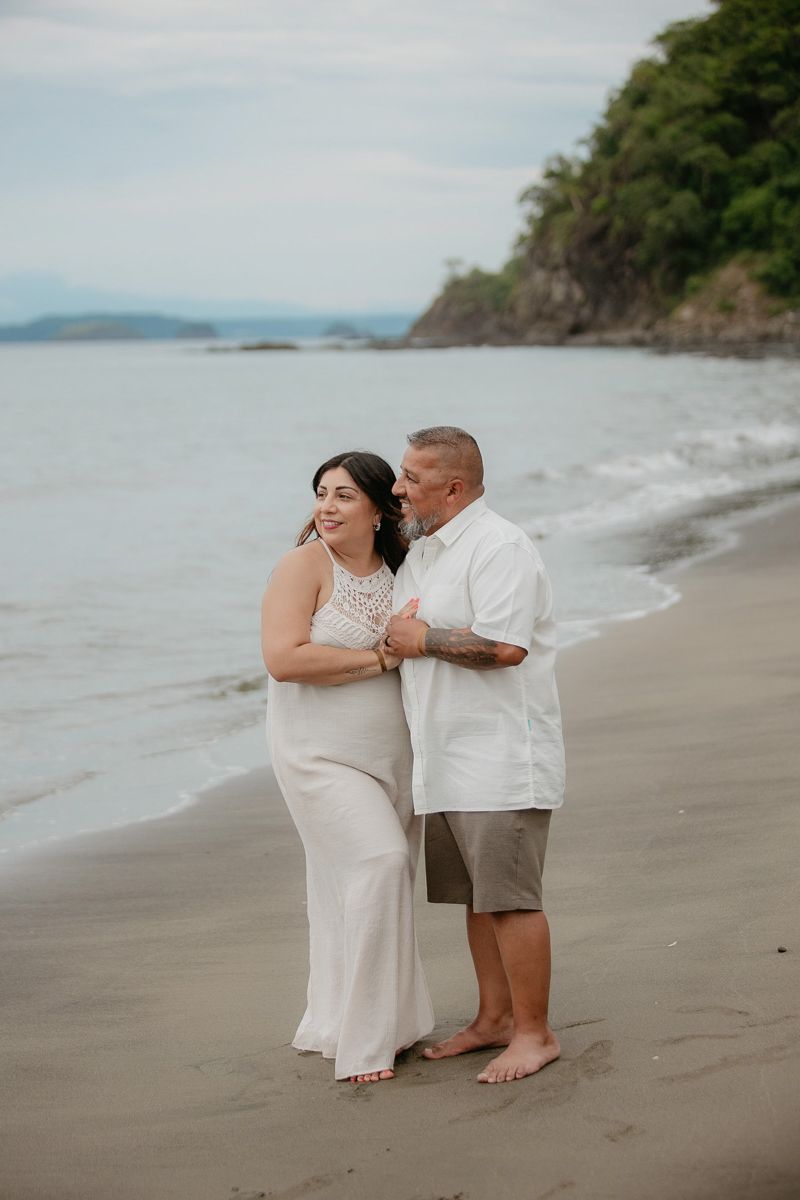 A couple shares an intimate moment while walking barefoot along a scenic beach with cliffs in the background.