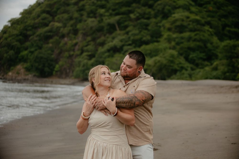 A couple embraces and laughs together on a secluded beach with lush green hills in the background.