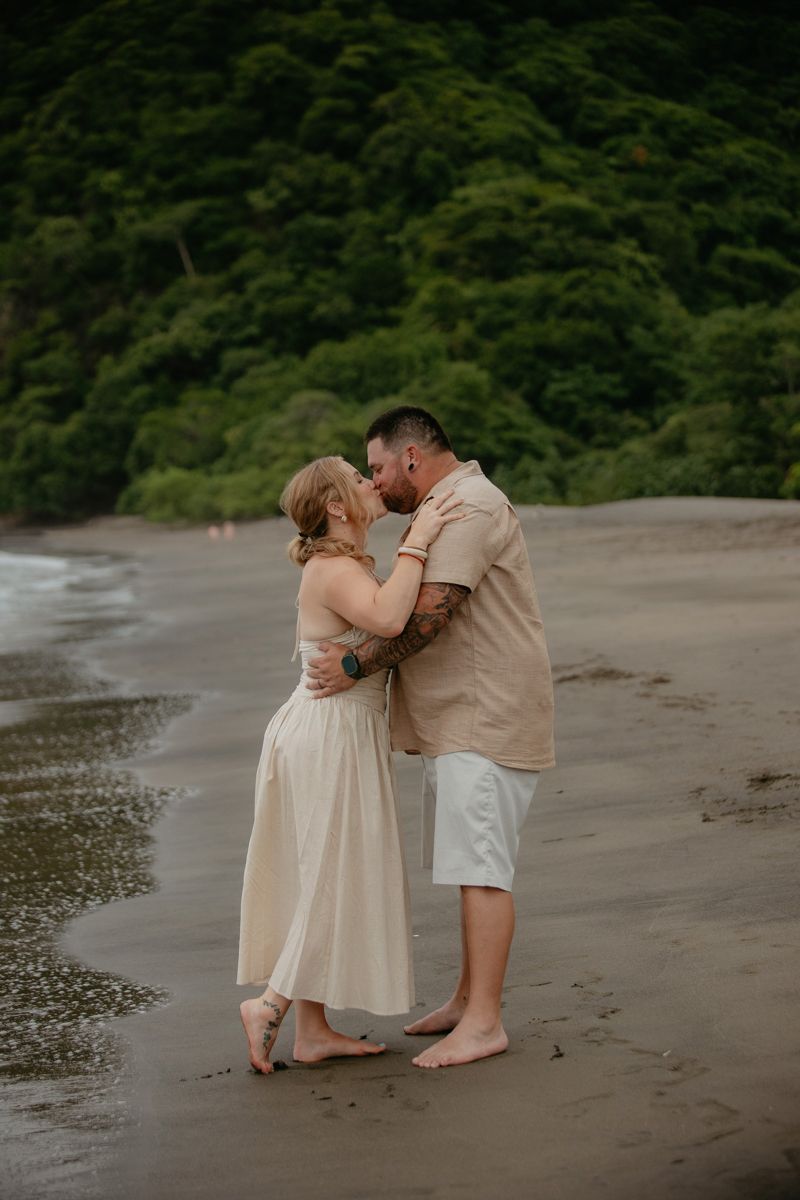 A couple sharing an intimate moment together on a dark sandy beach with lush greenery in the background.