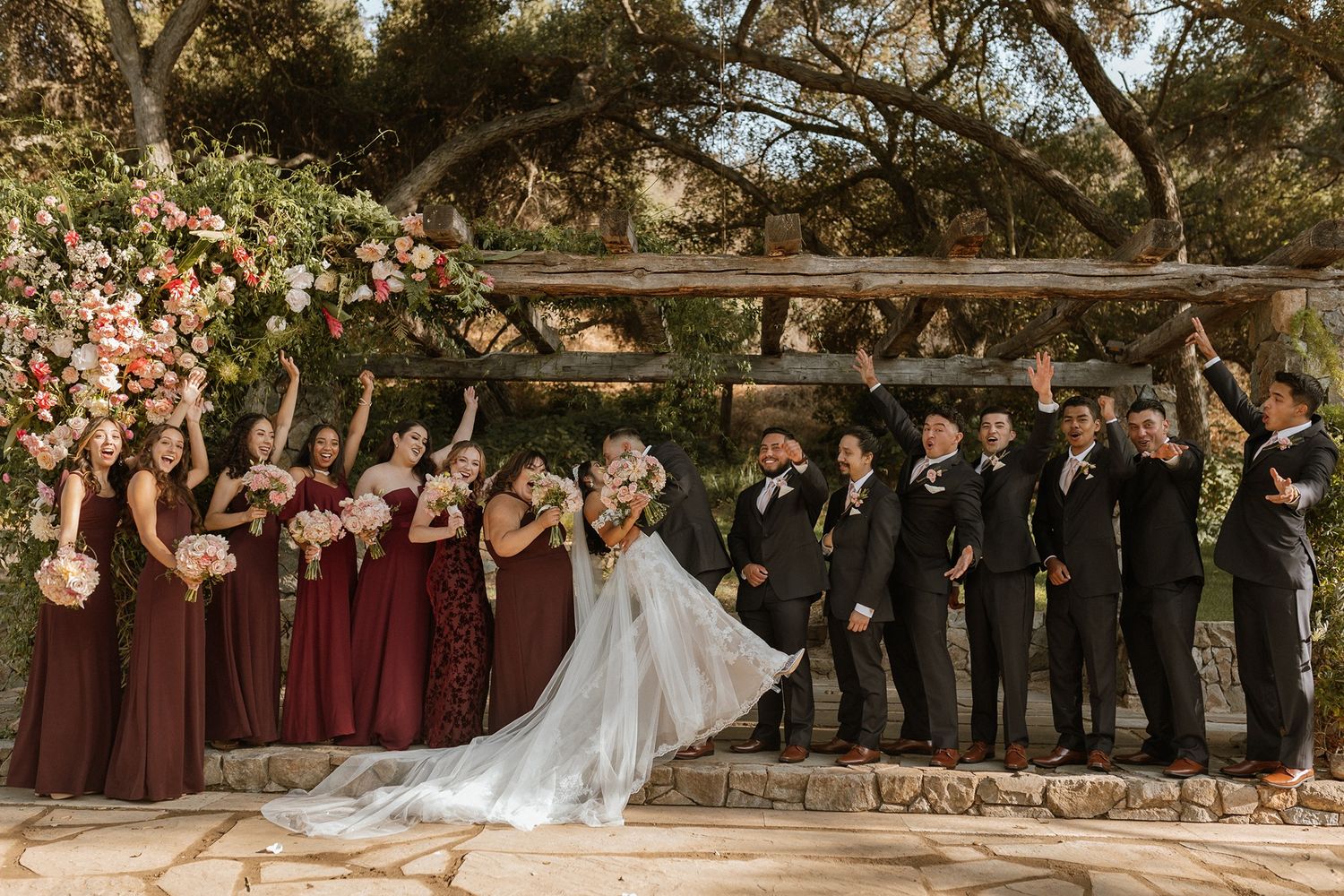 Wedding party celebrating under floral trellis, bridesmaids in burgundy dresses, groomsmen in black suits, bride and groom kissing.
