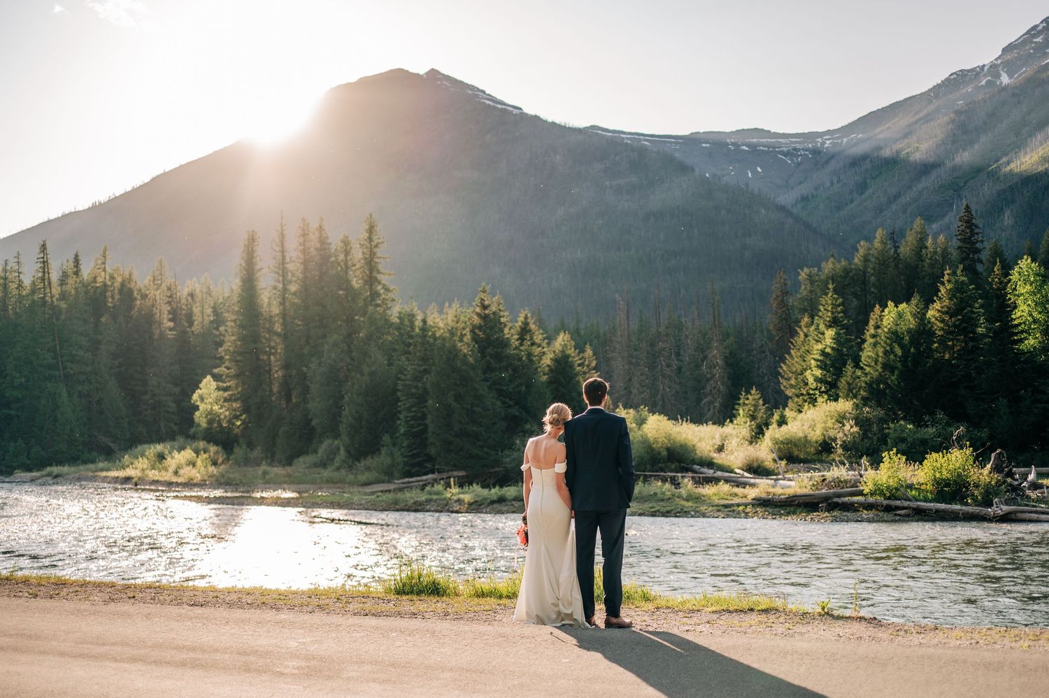 Bride and groom stand hand in hand by a mountain river, surrounded by pine trees and a glowing sunset backdrop.