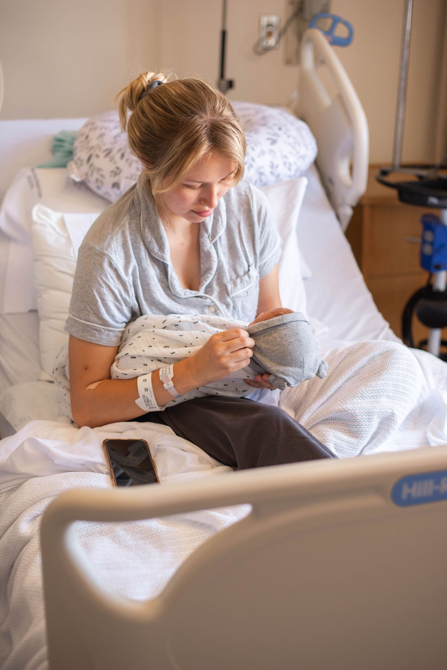New mother in hospital bed cradling her newborn baby, wearing a gray gown with an IV bracelet.