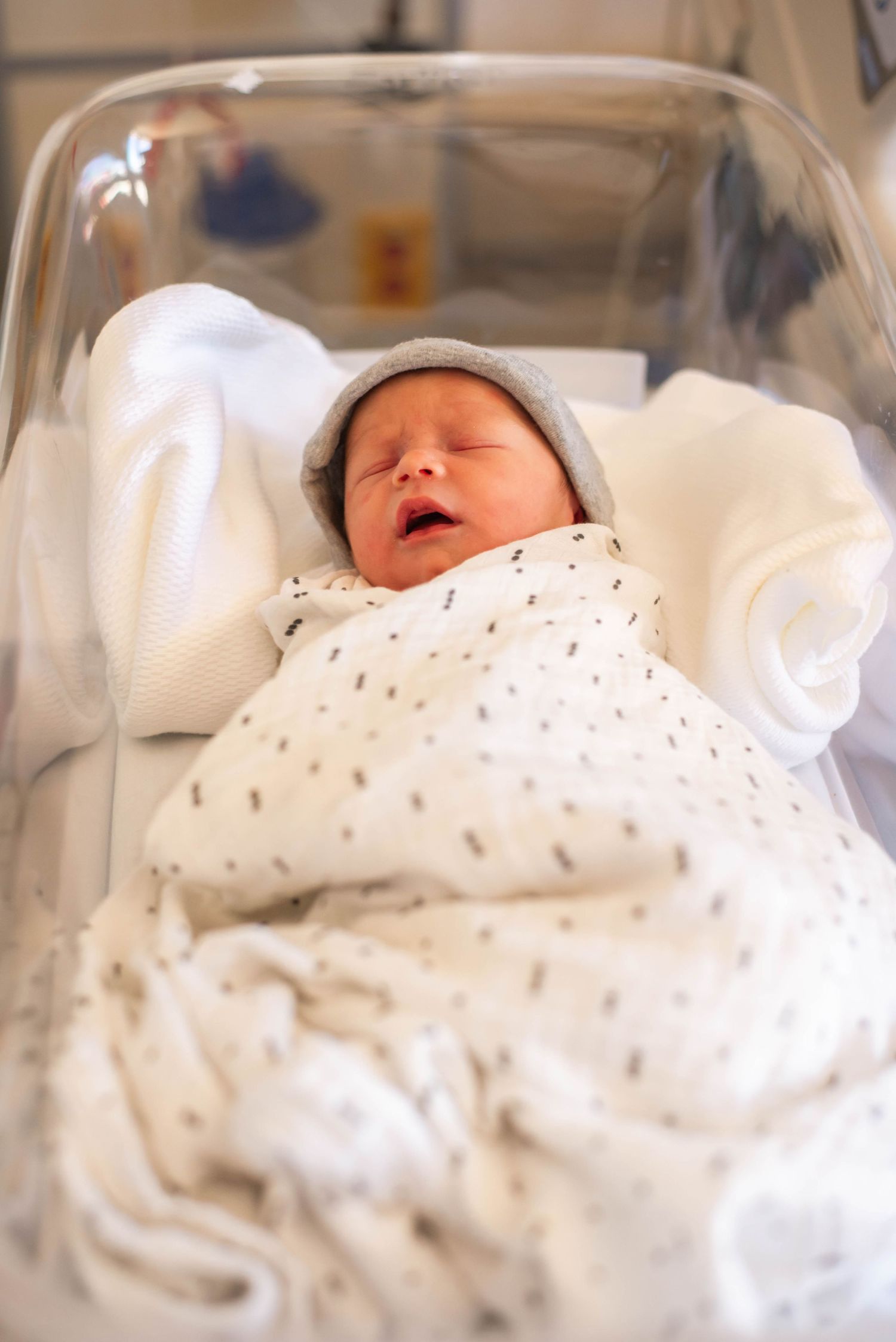 Peaceful newborn infant wrapped in white swaddle blanket with gray hat resting in clear hospital bassinet.