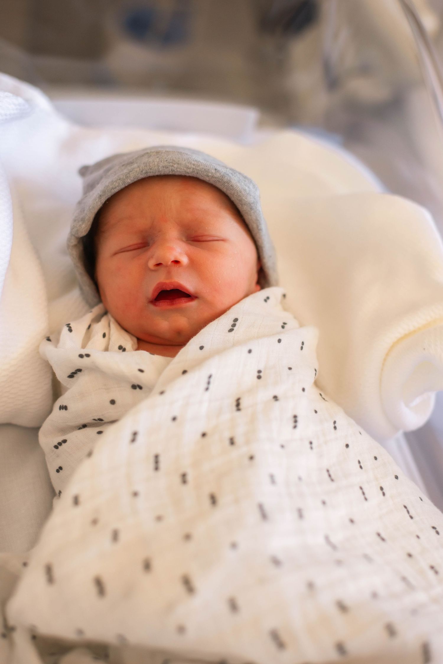 Newborn baby swaddled in white muslin blanket wearing gray knit hat sleeping in hospital bassinet.