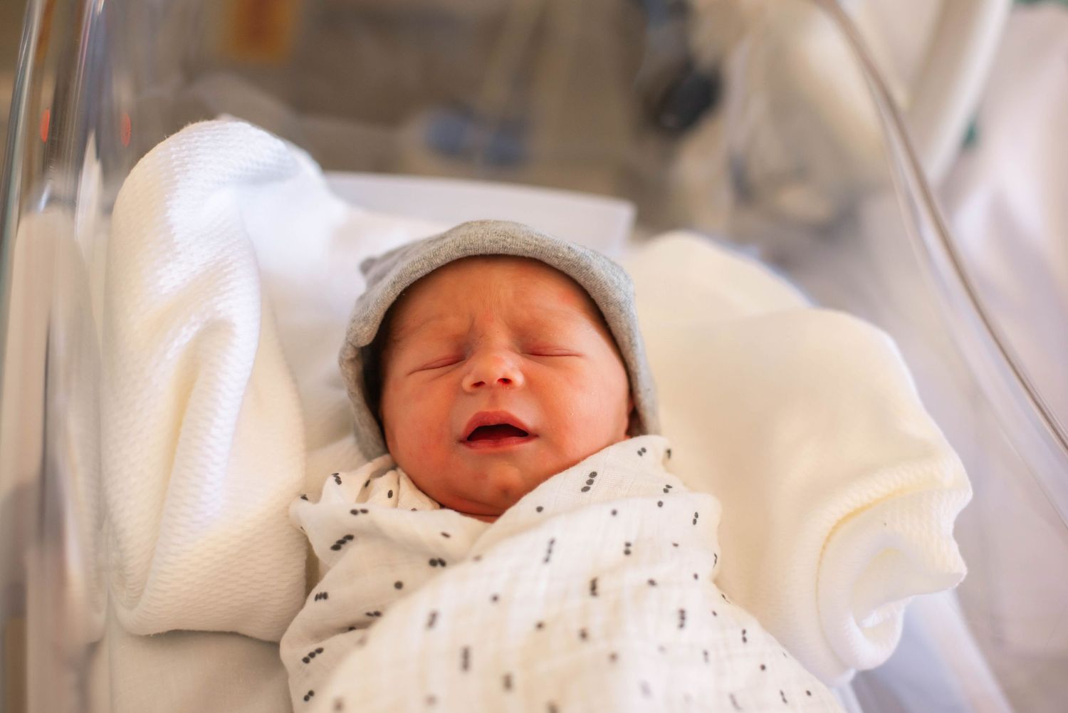 Newborn baby swaddled in white blanket wearing gray hat sleeping peacefully in hospital bassinet.