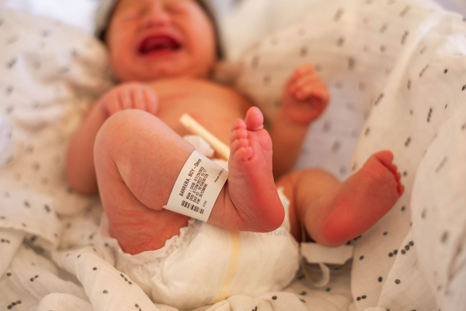 Close-up of crying newborn baby feet with hospital ID bracelet lying on white patterned blanket.