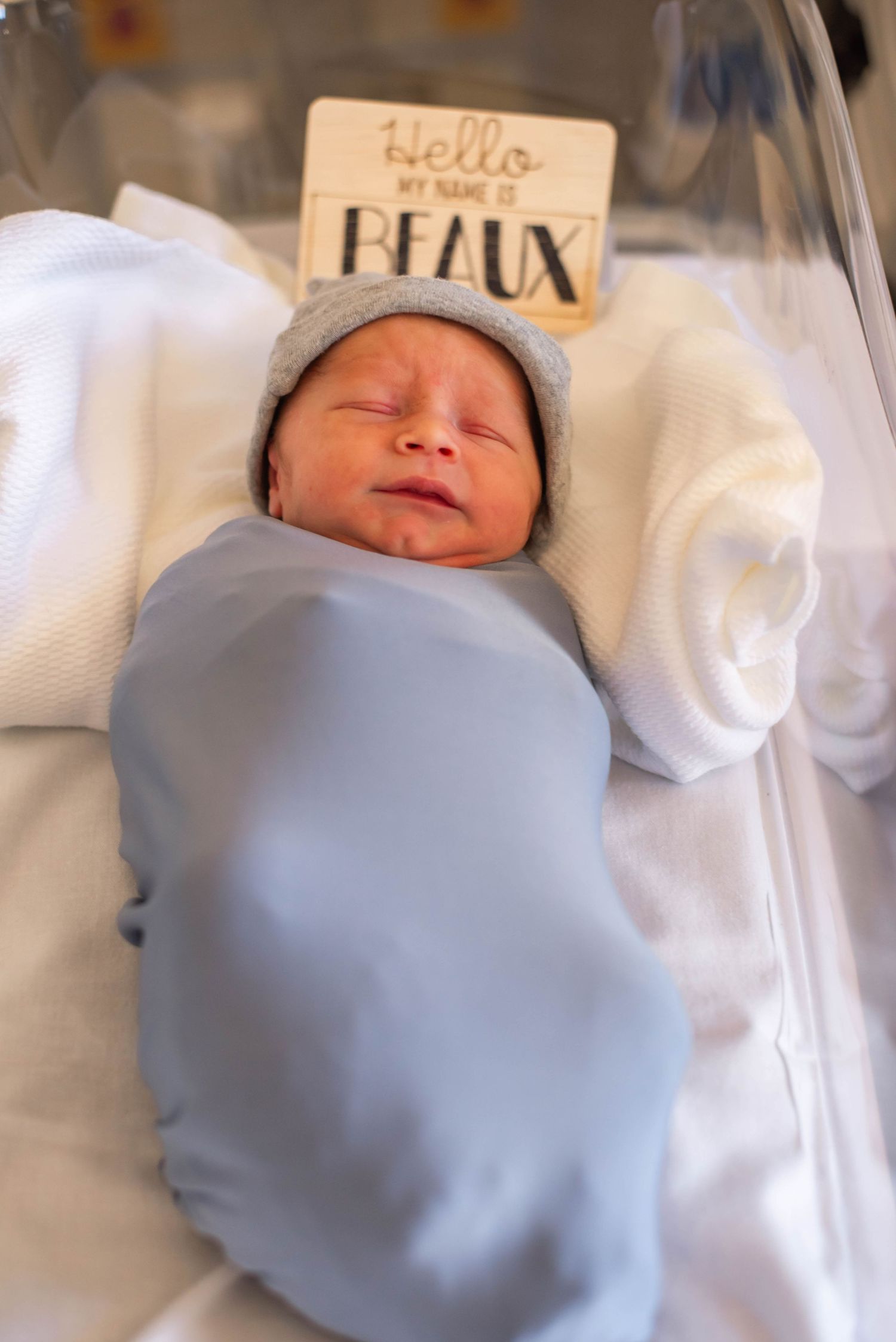 Newborn baby Beaux swaddled in blue blanket wearing gray hat in hospital bassinet with name sign.