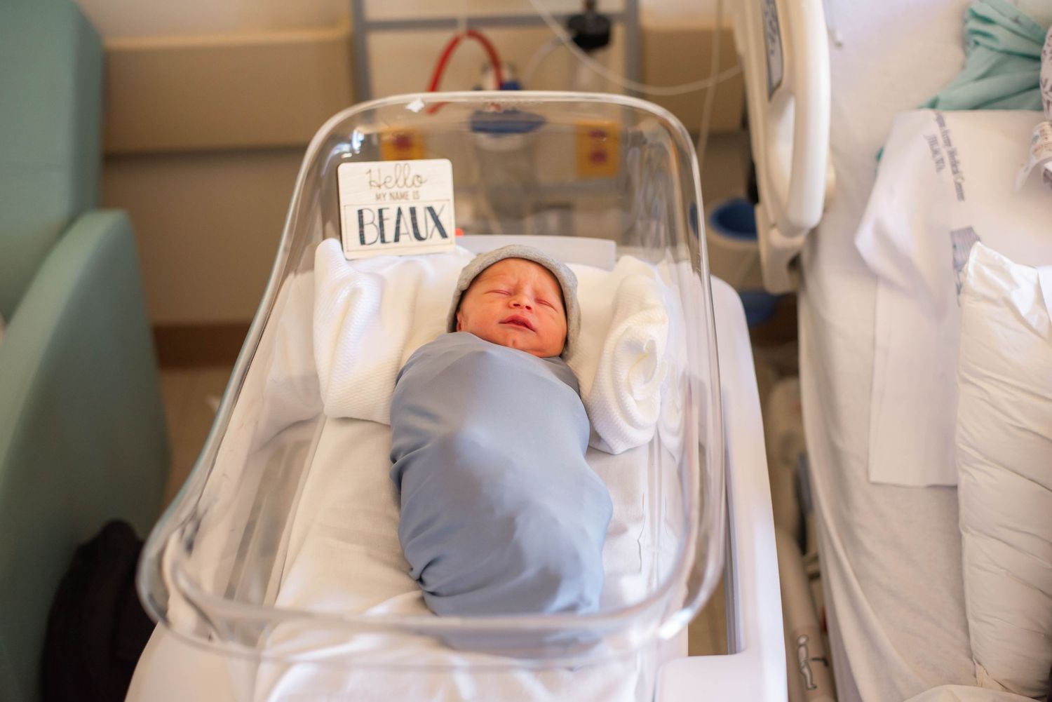 Swaddled newborn baby resting in clear hospital bassinet with name card.