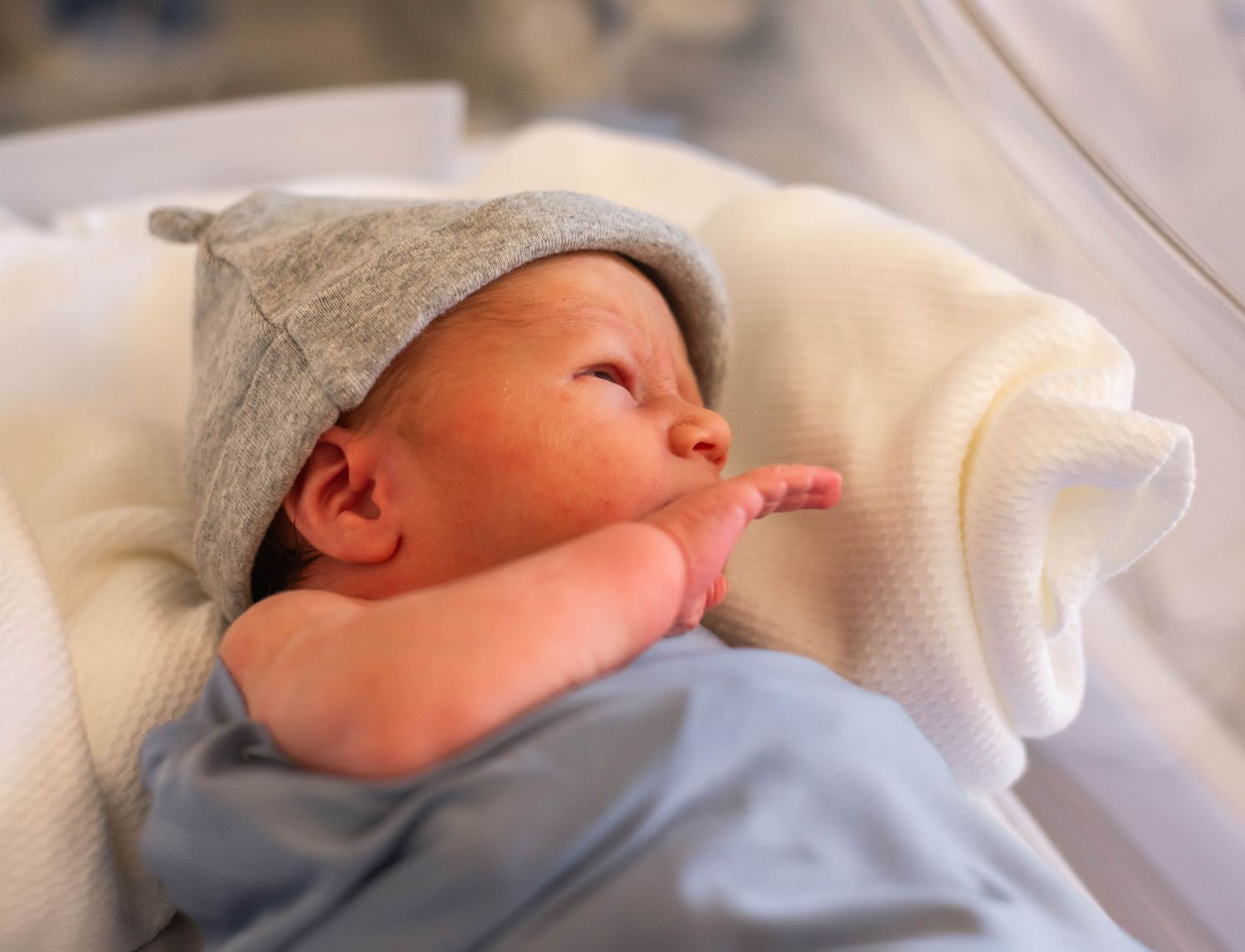 Newborn baby wearing gray knit hat lying on white blanket in hospital.