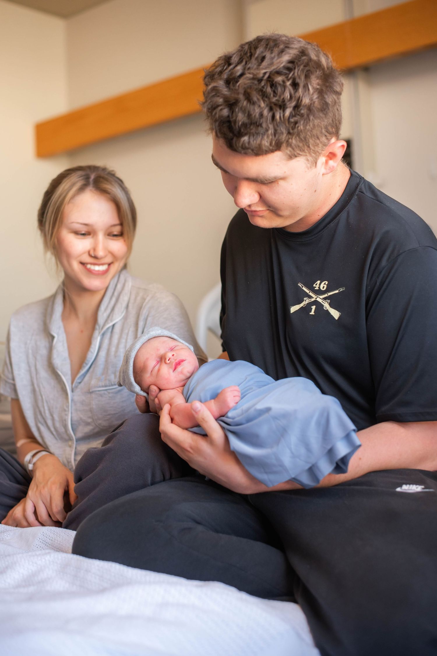 New parents smiling and holding their swaddled newborn baby in a blue blanket at the hospital.