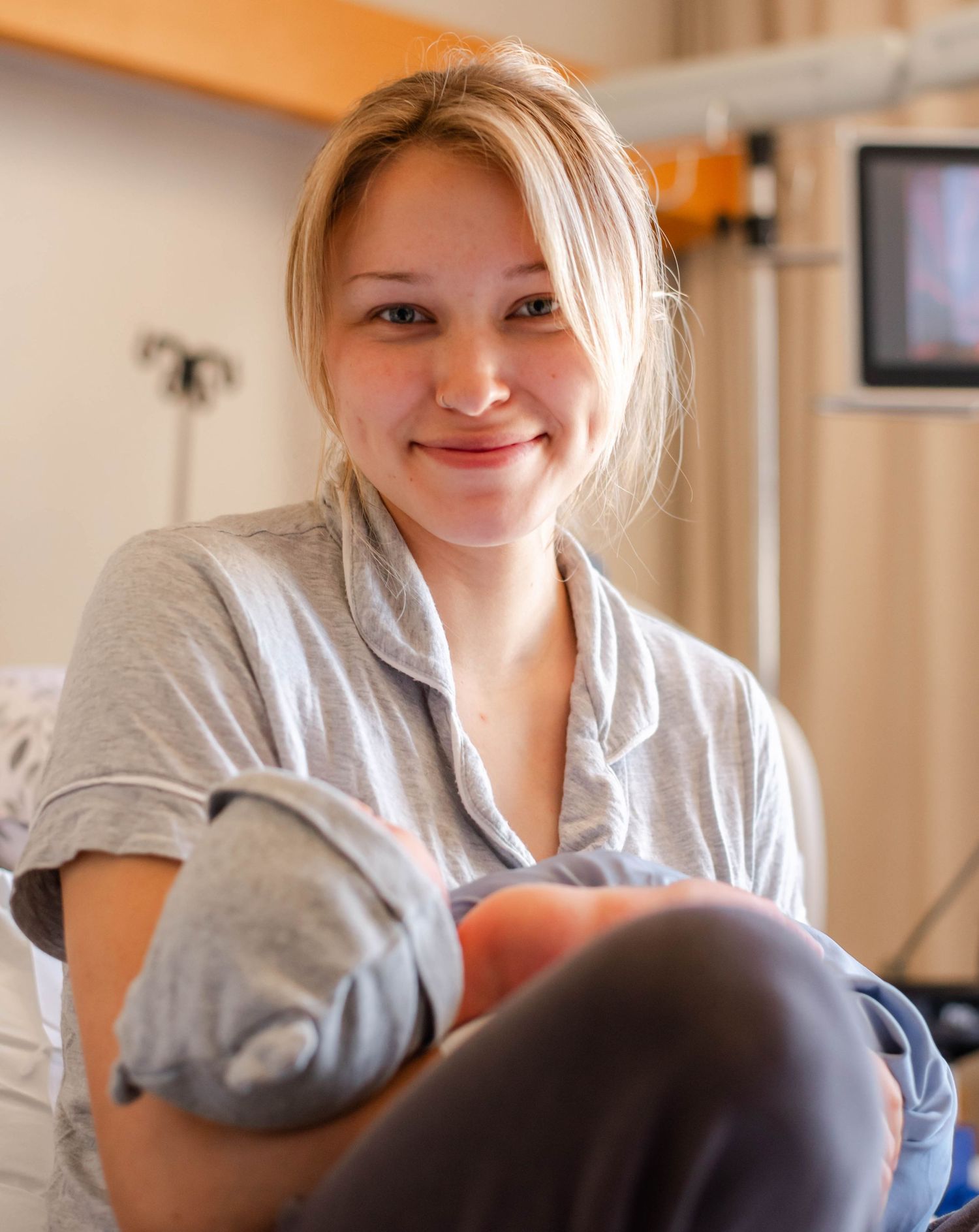 Smiling new mother in gray pajamas holding her newborn baby in hospital room after delivery.