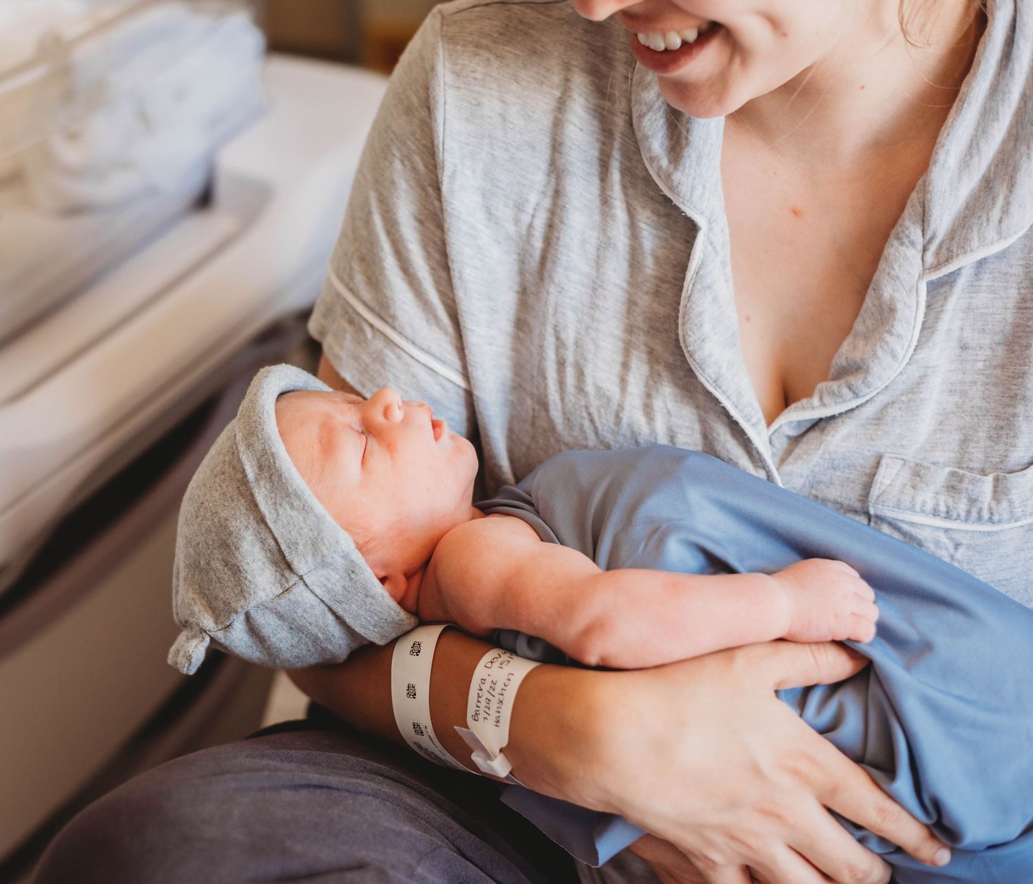 A smiling new mother holds her sleeping newborn baby in a hospital room, both wearing matching gray outfits.