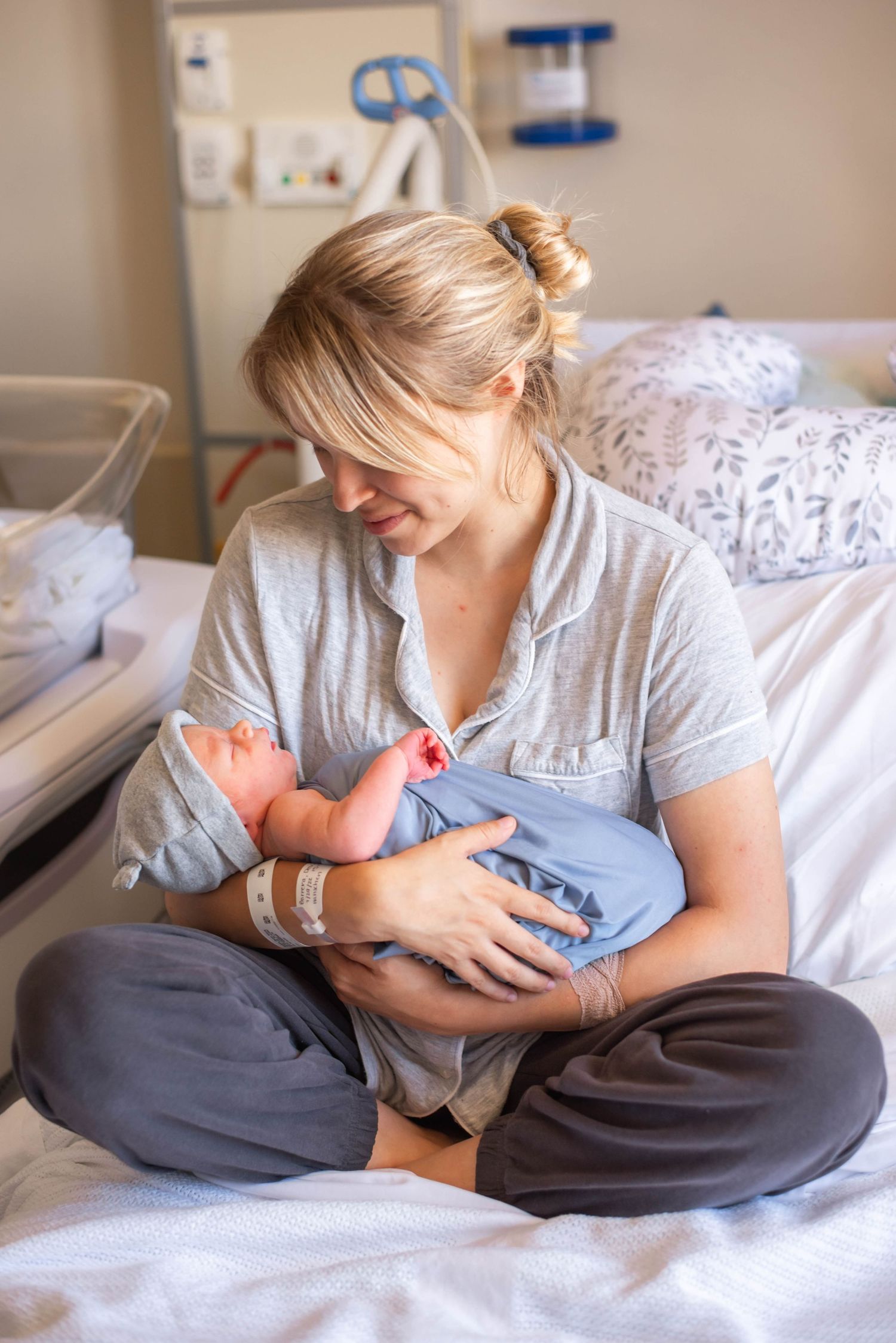 New mother in gray pajamas sitting cross-legged on hospital bed lovingly gazing at newborn baby.