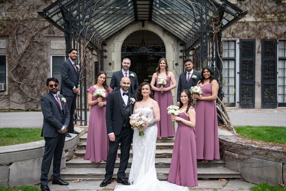 Wedding party poses on stone steps of ivy-covered mansion, bridesmaids in dusty rose gowns surrounding bride and groom in elegant attire.