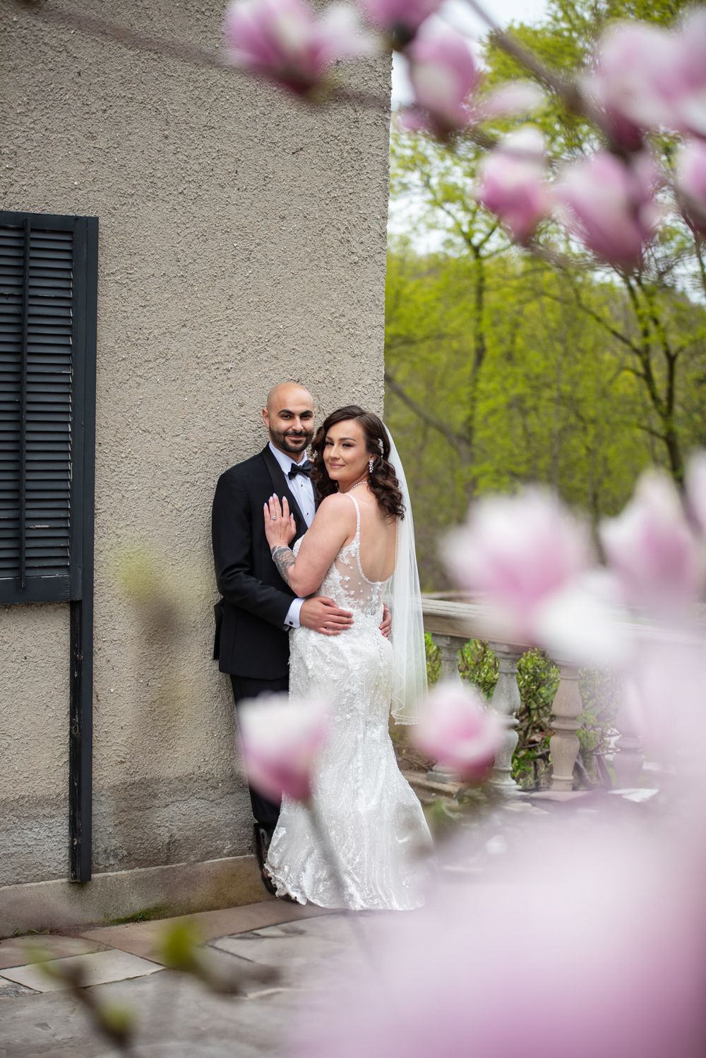 Bride and groom embracing against stone wall, framed by pink magnolia blossoms in foreground.