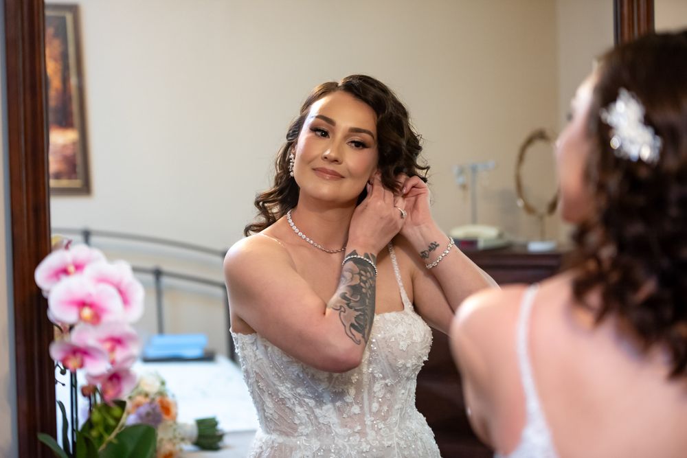 Bride with dark hair and tattoos adjusts her necklace while getting ready in front of a mirror.