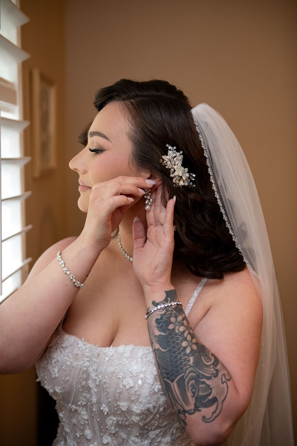Bride in lace gown puts on earrings with floral hair comb and veil by a window.