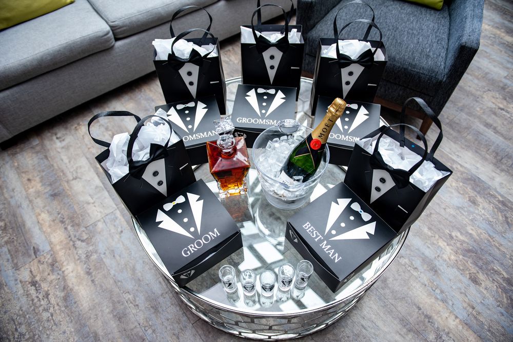 Tuxedo-themed groomsmen gift bags arranged around champagne and whiskey on a mirrored table.