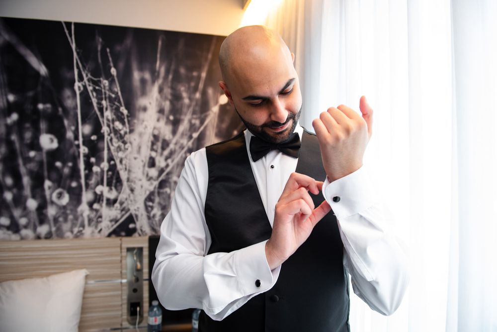 Dapper bald groom in black vest and bow tie adjusts his cufflinks while getting ready.