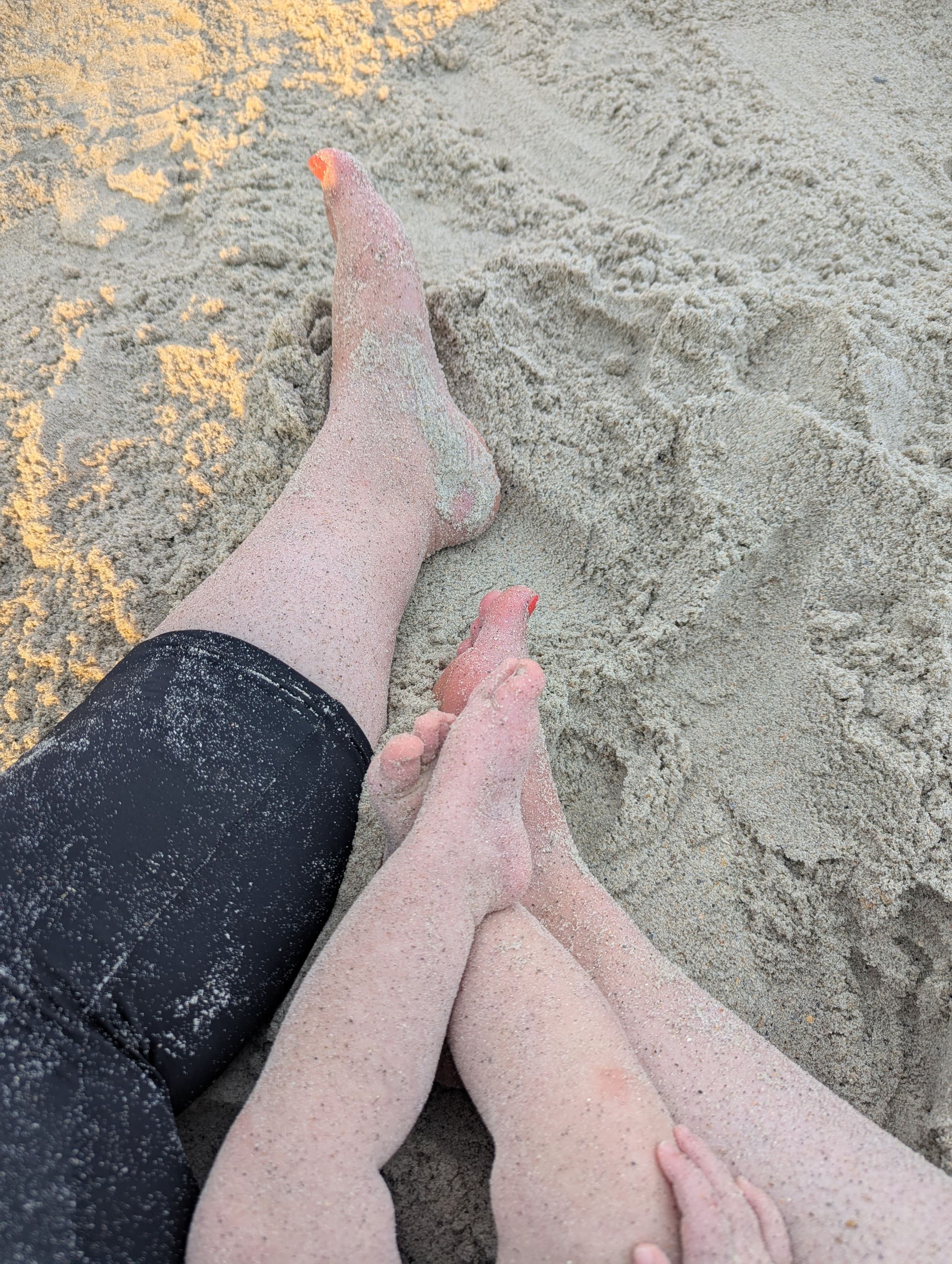 Two people sitting on sandy beach with legs stretched out, one with orange toenail polish, sand covering their feet.