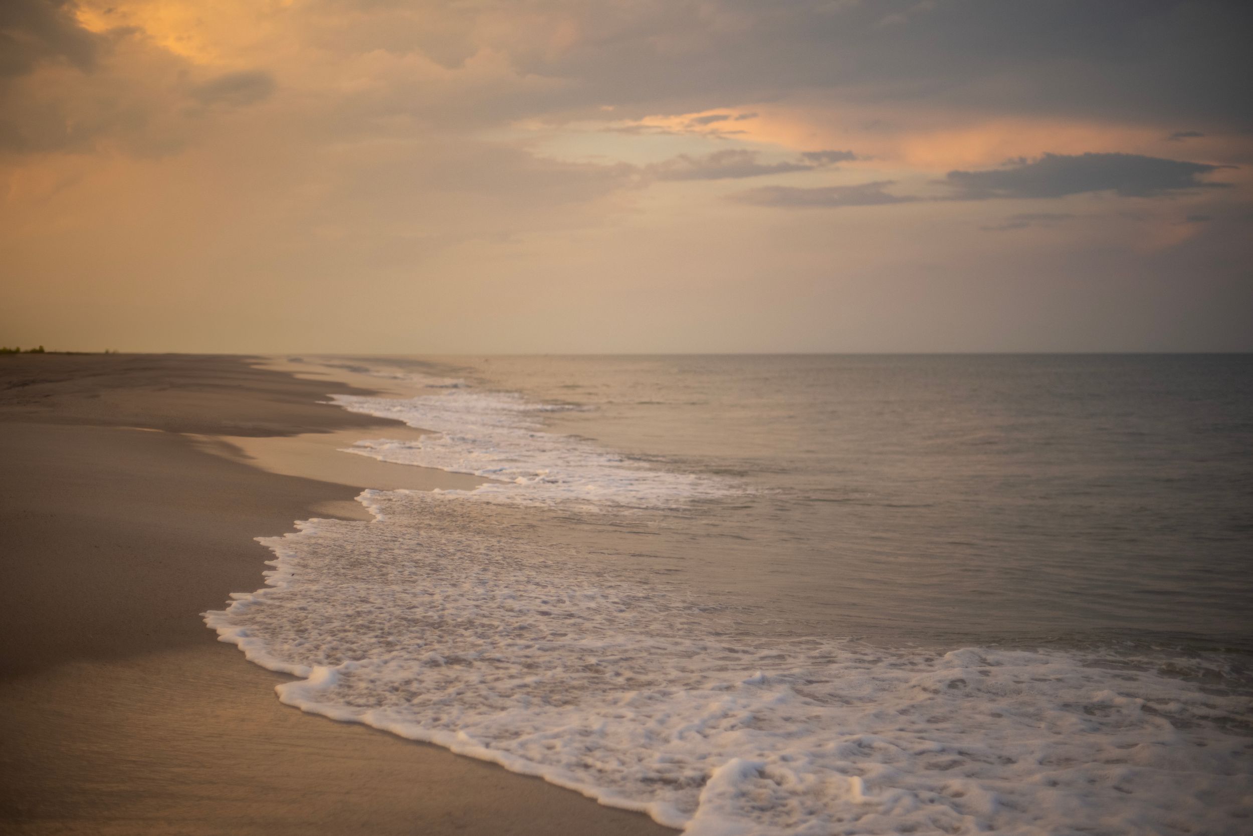 Serene beach at sunset with gentle ocean waves washing ashore on golden sand under a warm, cloudy sky.