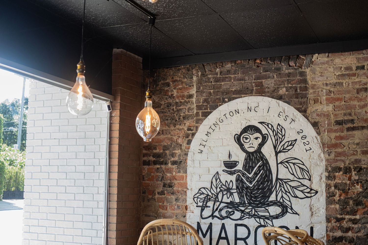 Cozy cafe interior with exposed brick wall, monkey mural, Edison bulb lights, and wicker chairs at Marcel's in Wilmington, NC.