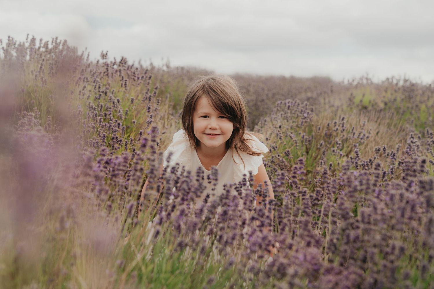 Young girl smiling in a blooming lavender field, surrounded by purple flowers on a cloudy day.