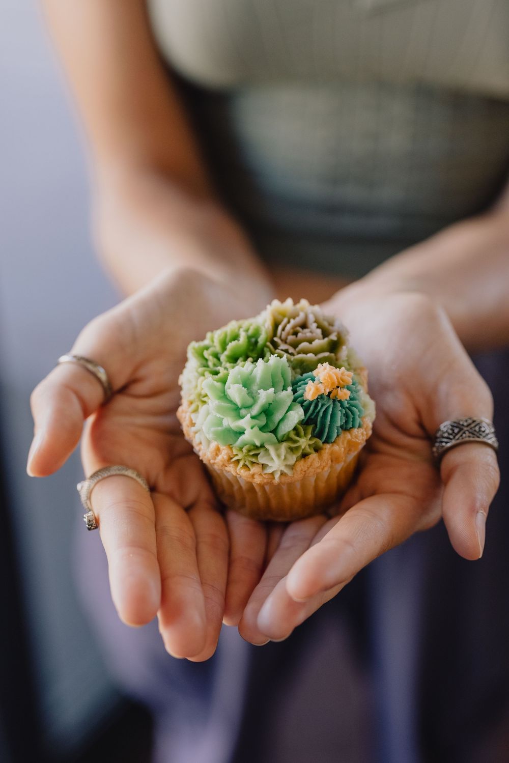 Hands holding a decorative succulent-themed wedding cupcake with green buttercream frosting.