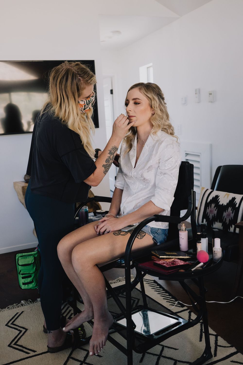 Makeup artist applying bridal makeup to a blonde bride seated in a chair during wedding preparations.