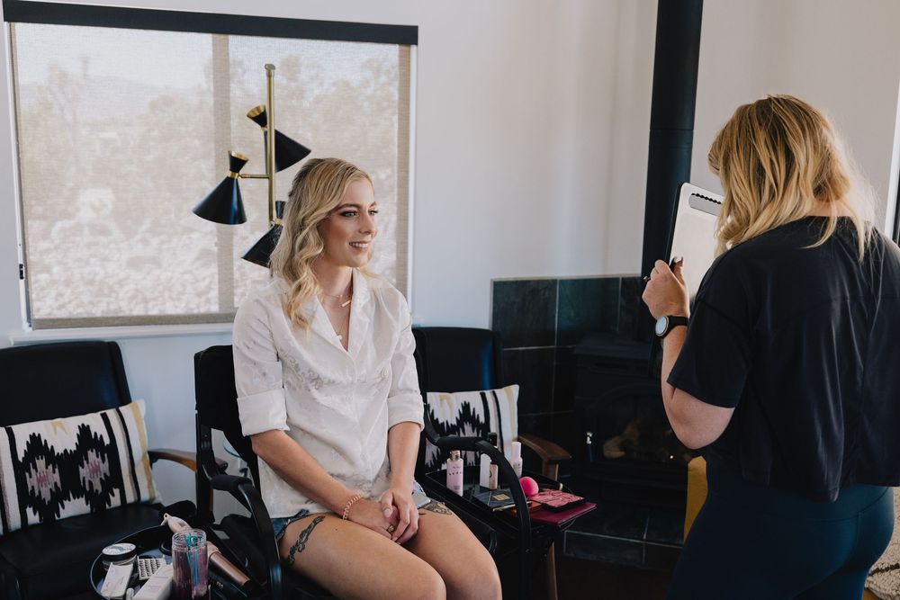 Blonde bride smiles during makeup application in a bright modern room before her wedding ceremony.