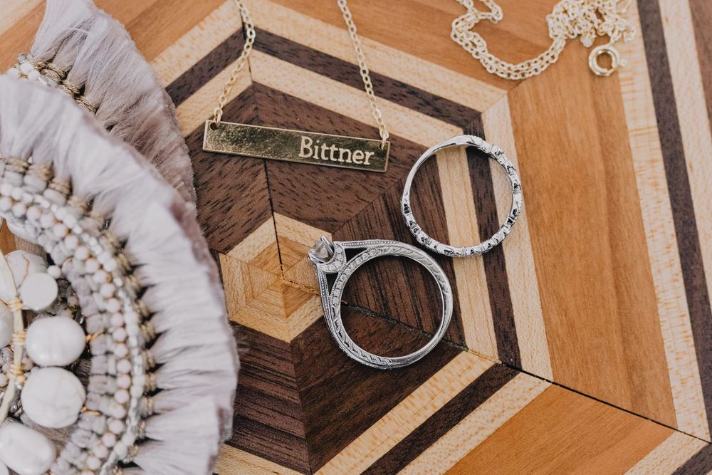 Wedding details flat lay featuring Bittner name bar necklace, diamond ring set, and boho accessories on wood surface.