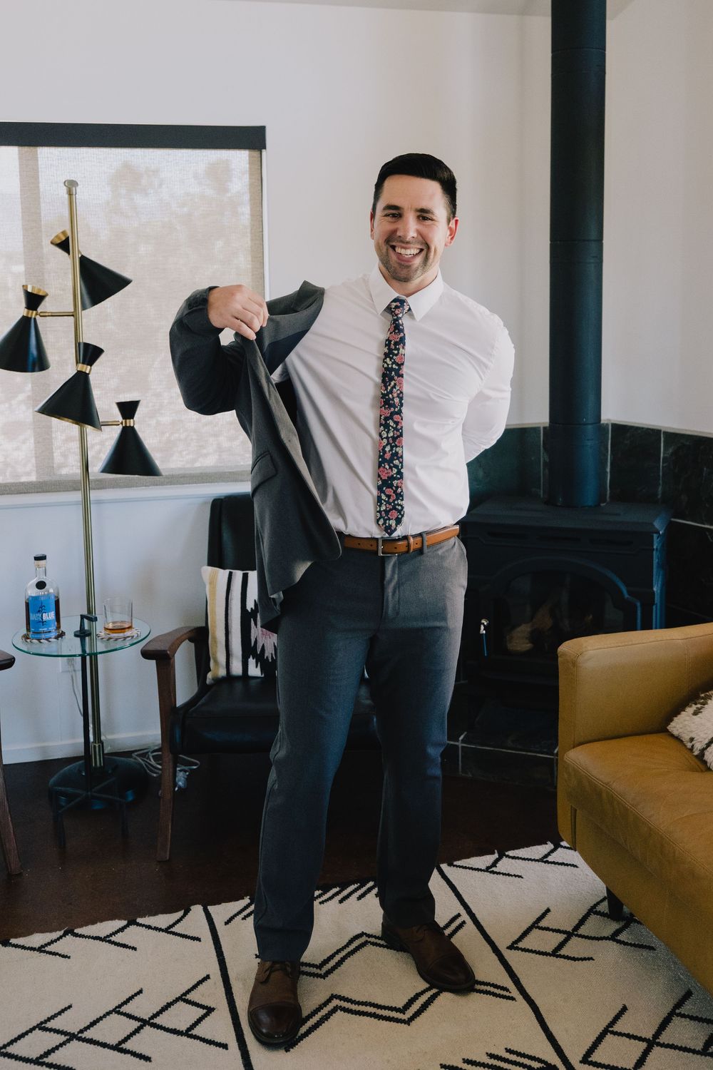 Smiling groom putting on dark suit jacket wearing floral tie in modern interior setting.