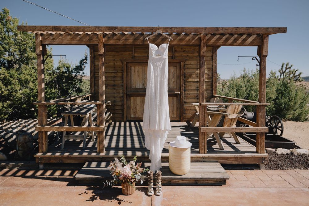 White wedding dress hanging on rustic wooden cabin porch with cowboy boots and wildflower bouquet below.