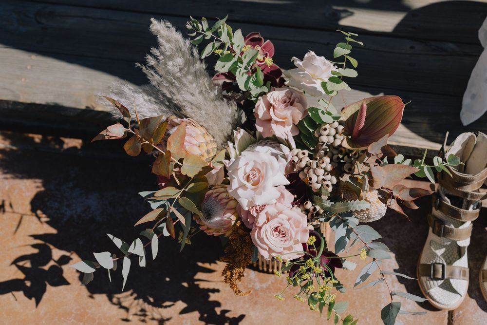 Rustic boho wedding bouquet with blush roses, pampas grass, and eucalyptus beside white strappy bridal heels outdoors.