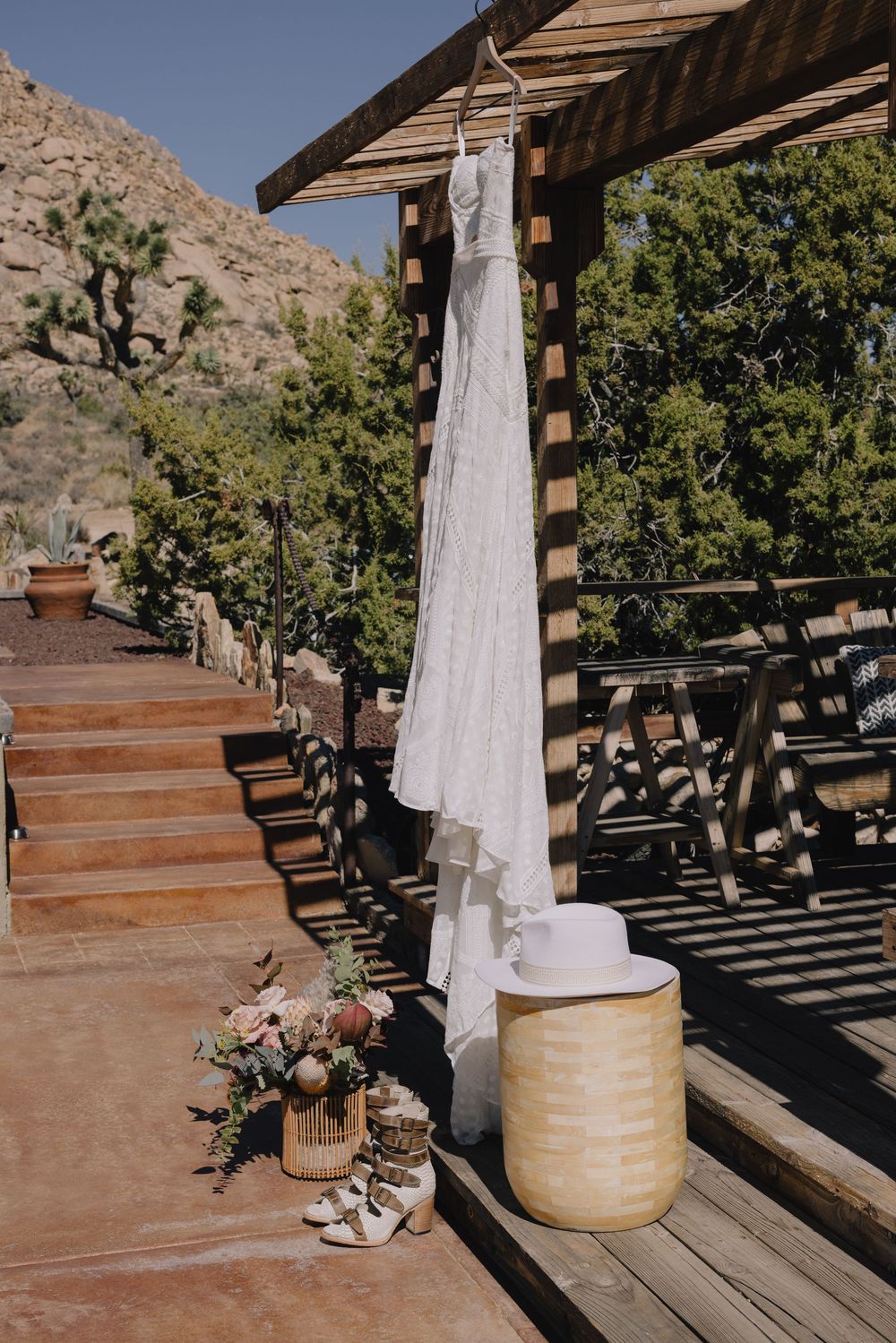 Boho wedding dress hanging on rustic wooden pergola with bridal accessories at desert venue.