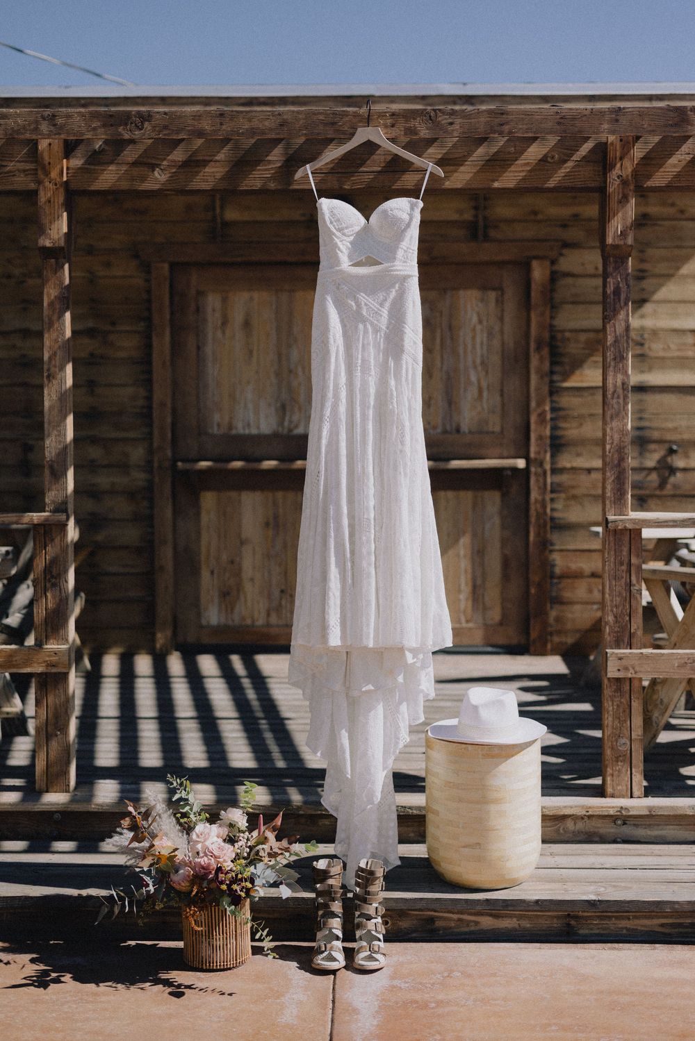 Strapless lace wedding dress hanging outside a rustic wooden cabin with bridal accessories below.