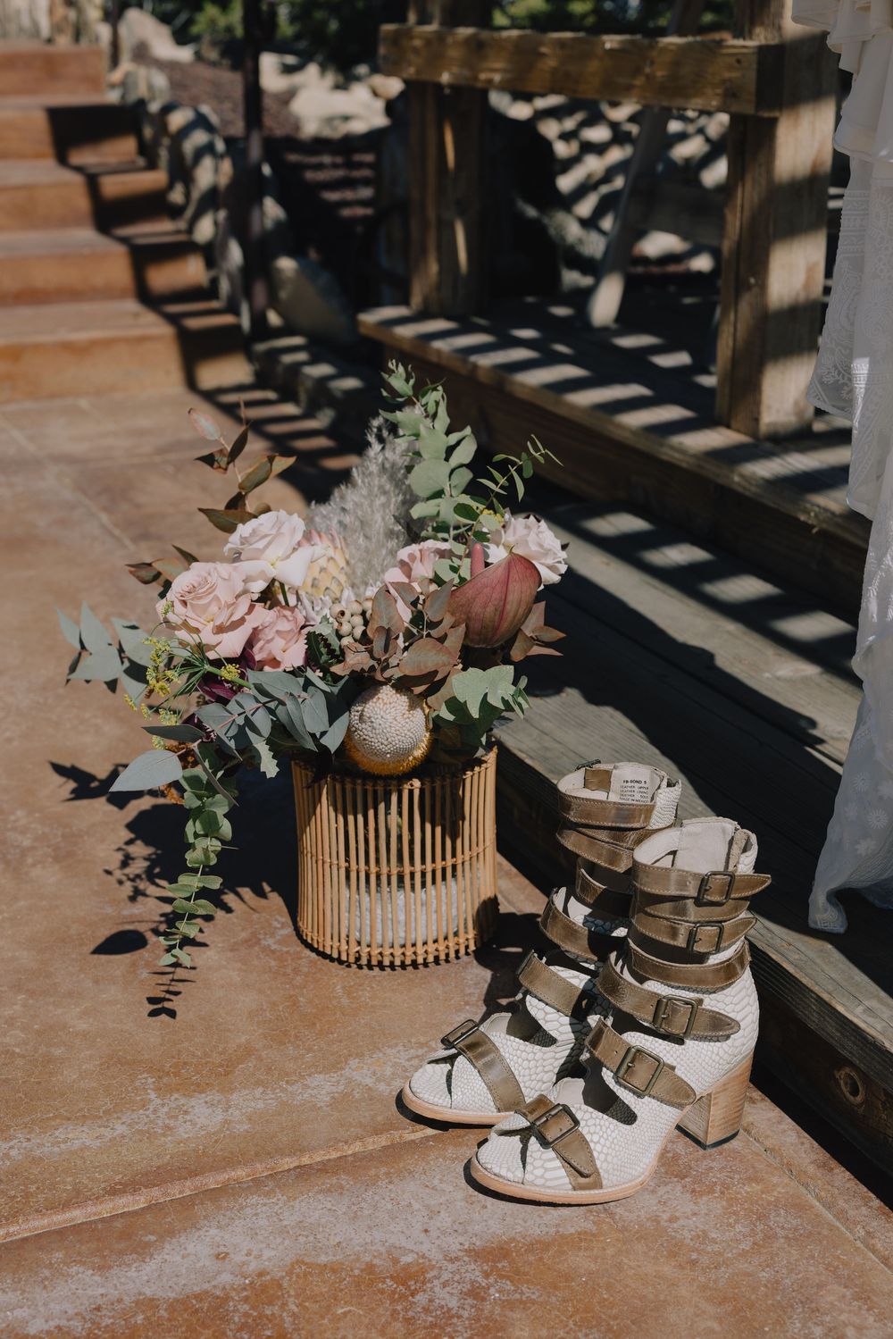 Boho wedding flat lay with blush floral arrangement in bamboo vase beside white strappy block heel bridal shoes.