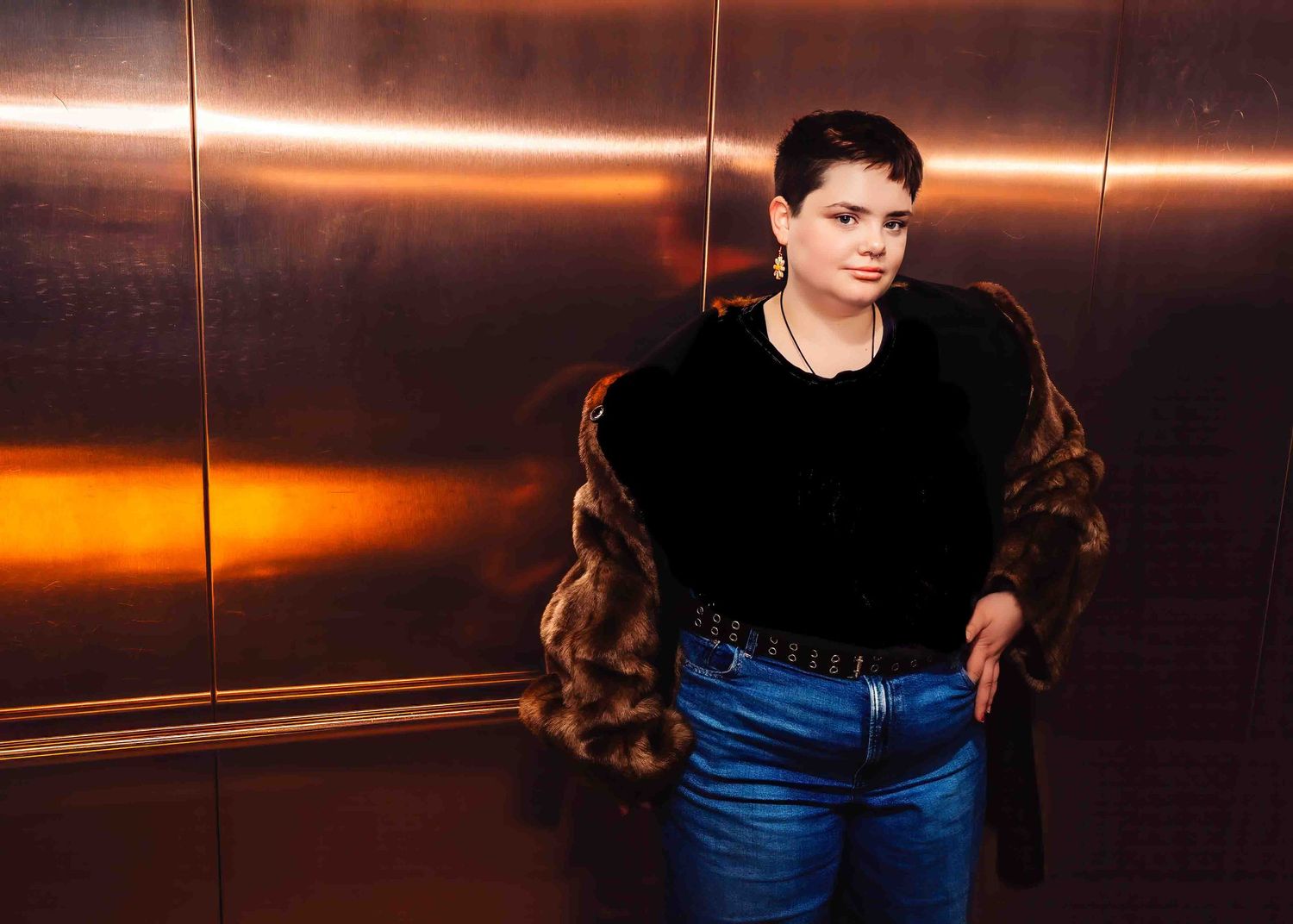 Woman with short hair wearing a fur coat and jeans, standing in a golden elevator.