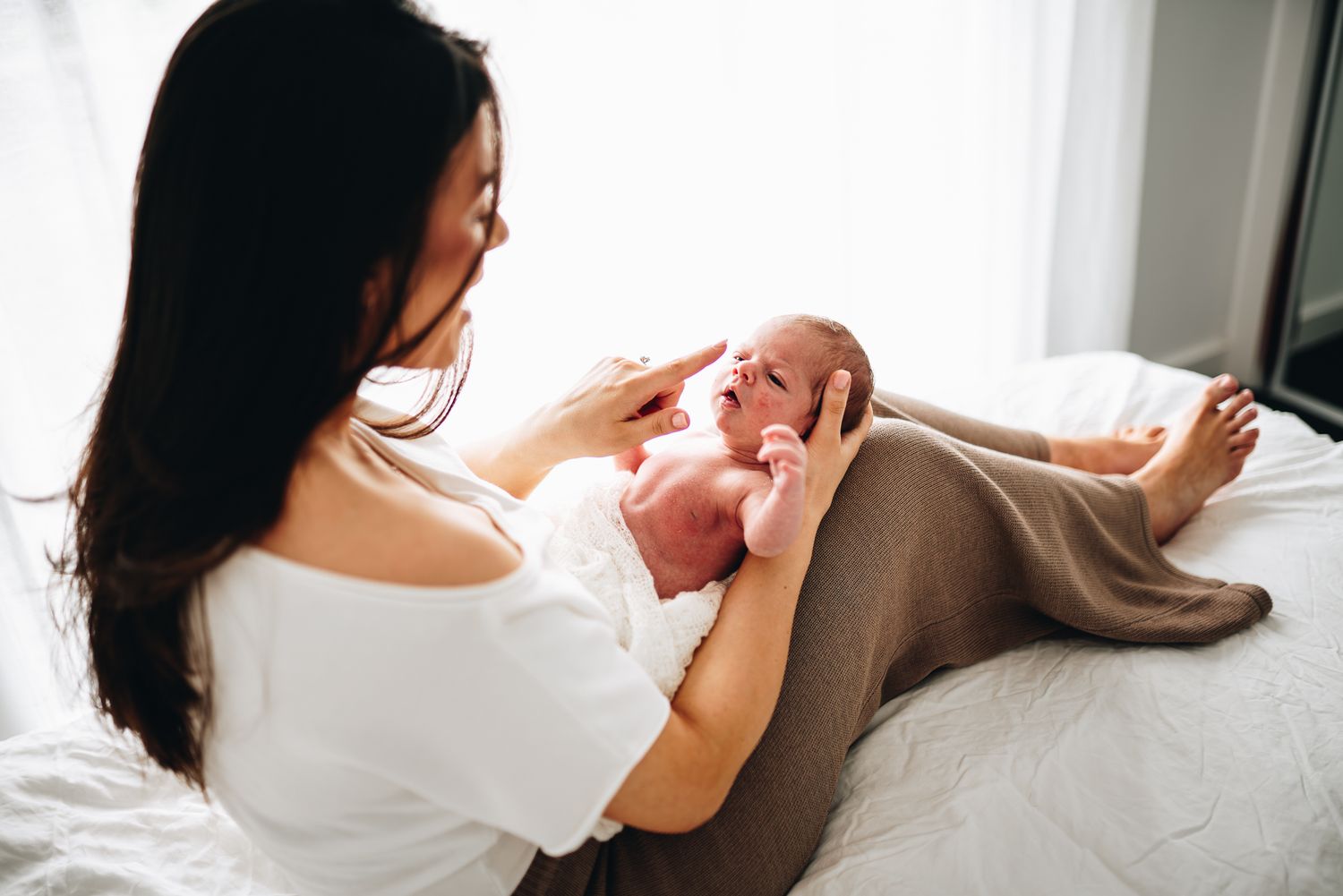 A mother tenderly holds her newborn baby on a white bed, gazing lovingly at her infant taken by a Melbourne newborn photographer.