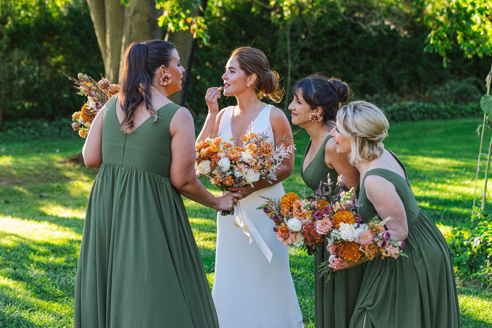 A bride in a white dress stands with her bridesmaids in green dresses, holding colorful bouquets, in a lush garden setting.