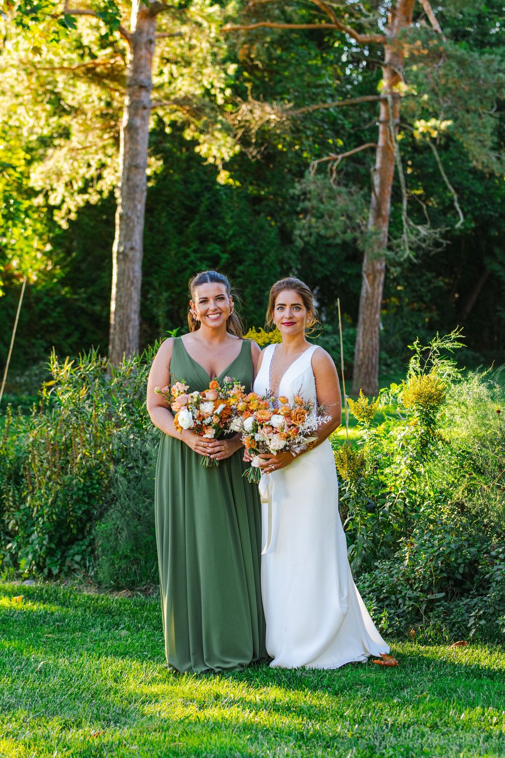 Two women in green and white dresses holding bouquets of flowers stand in a lush garden.