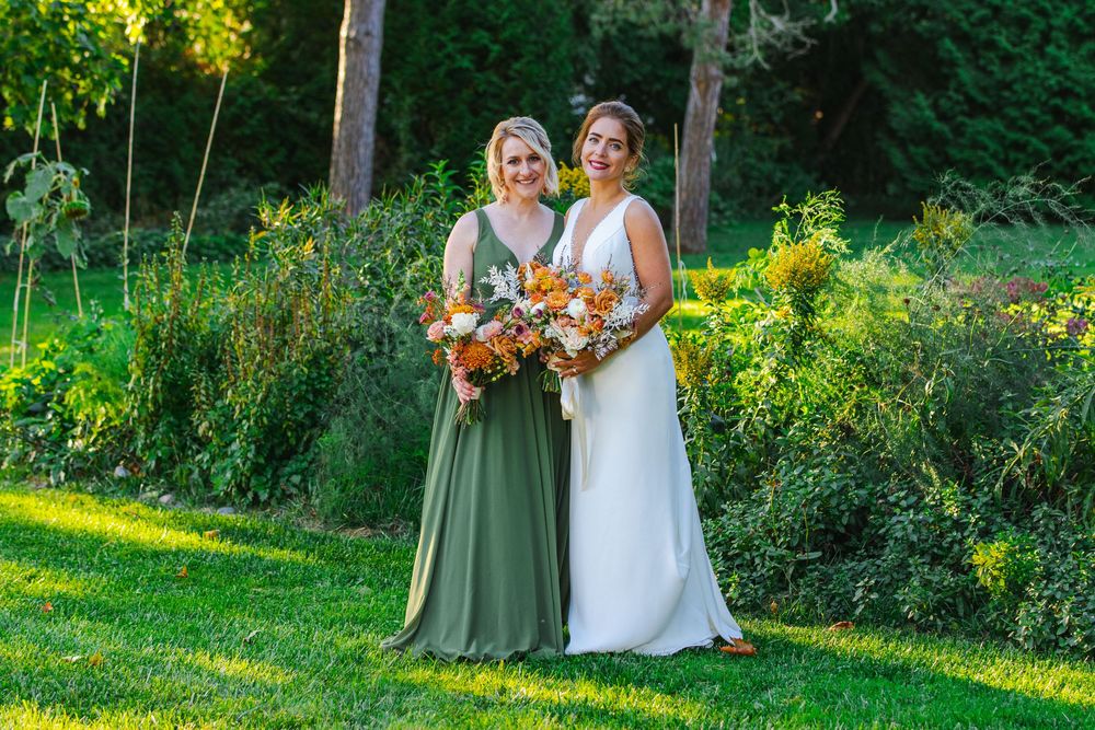 Two women in formal attire stand together in a garden, holding a bouquet of flowers.