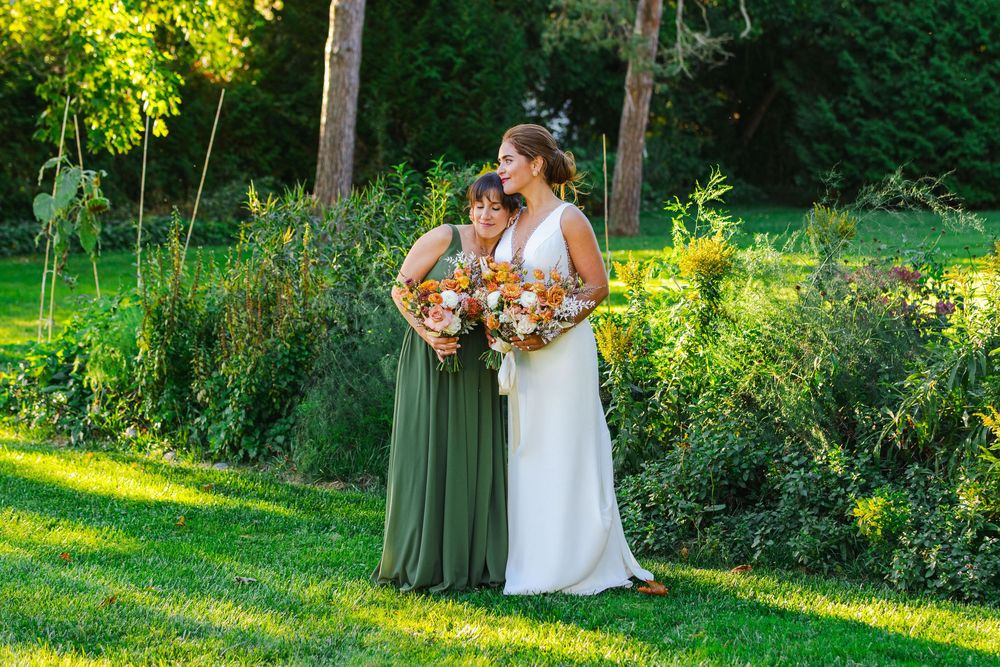 A bride in a white dress and a bridesmaid in a green dress hold bouquets of flowers in a garden.