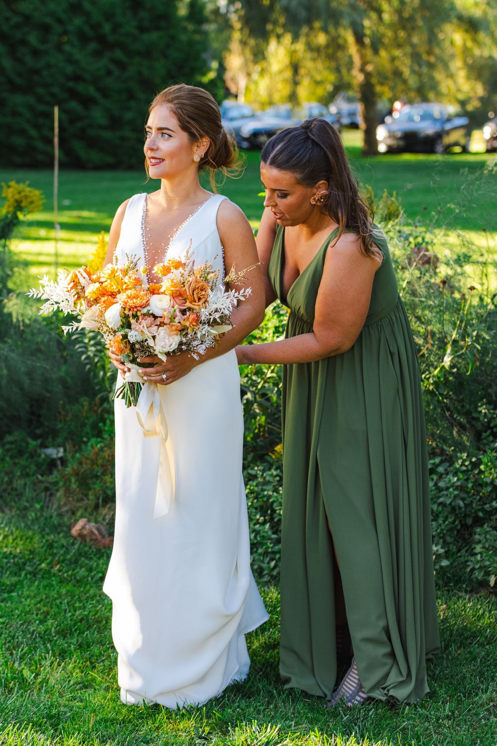 Two women in formal dresses stand together in a garden, holding bouquets of flowers.