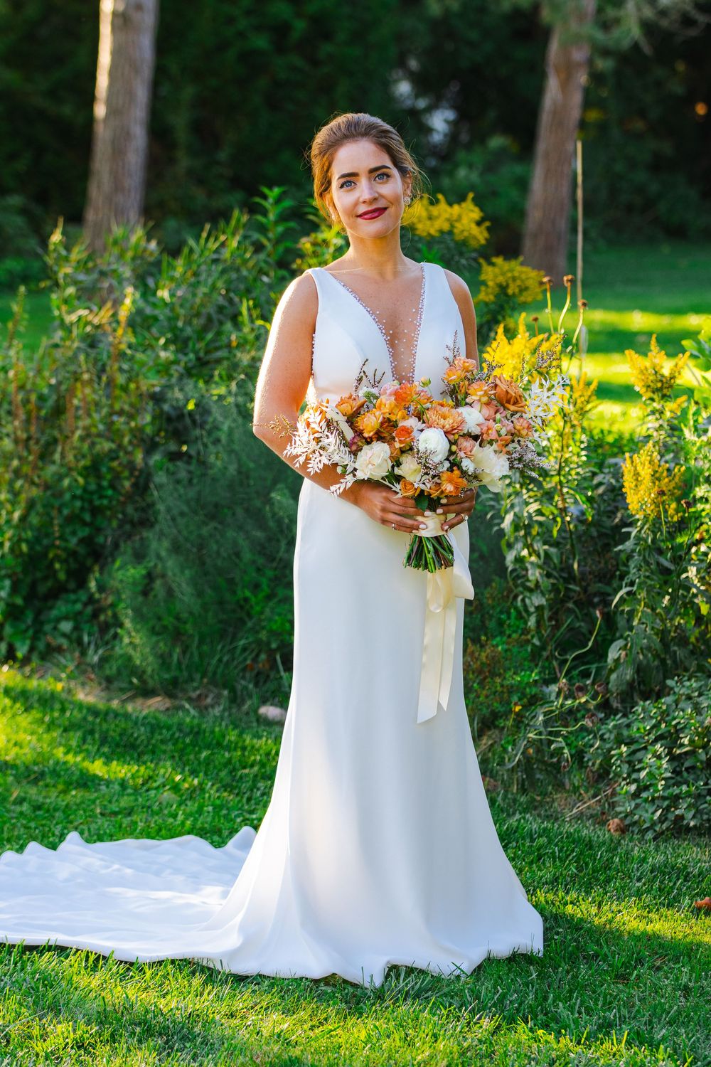 A woman wearing a white wedding dress stands in a garden holding a bouquet of colorful flowers.
