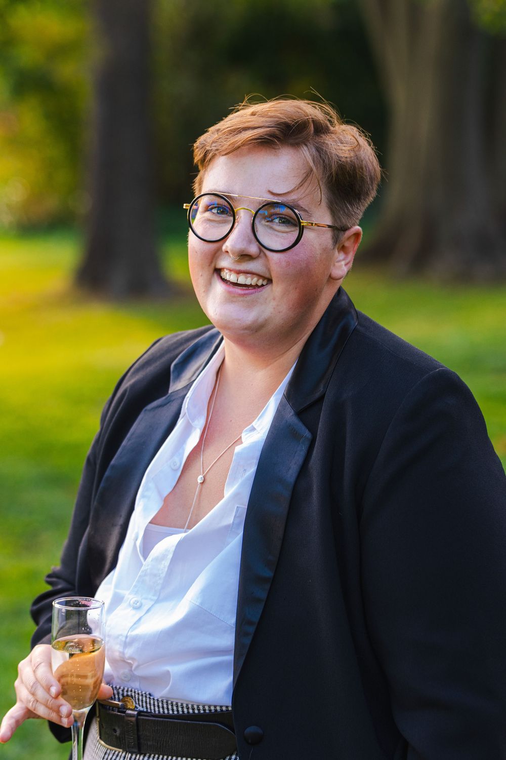 A smiling woman with glasses stands in a garden, holding a glass.