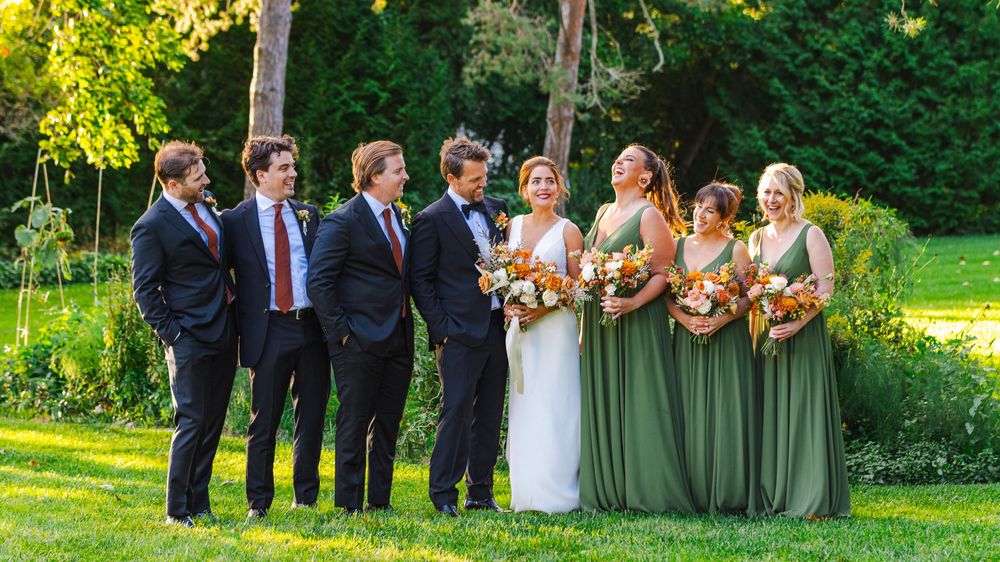A wedding party, including the bride in a white dress and bridesmaids in green dresses, stand together in a garden.