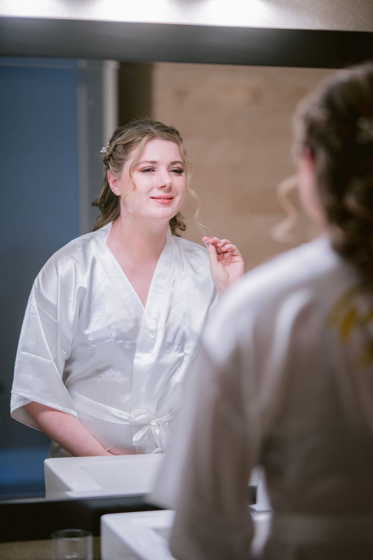 Smiling bride in white satin robe admiring her wedding hair and makeup in a bathroom mirror.
