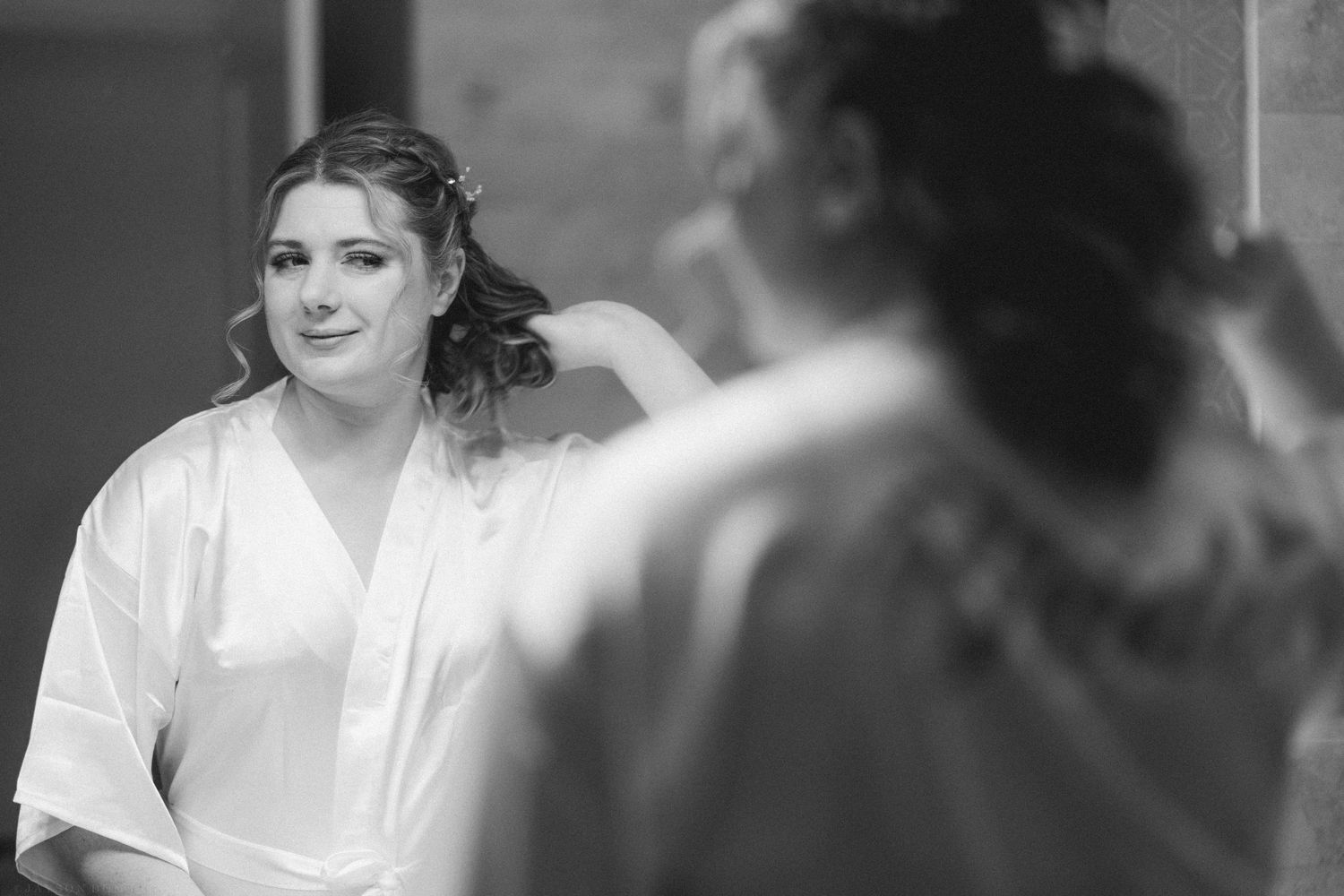 Black and white photo of a bride in a white robe getting hair done by a bridesmaid before her wedding.