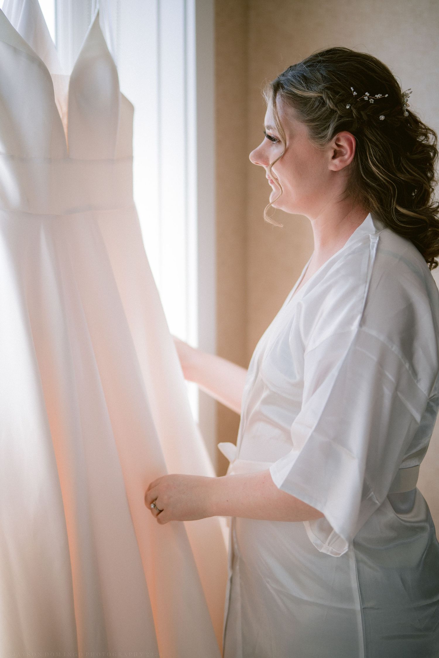 Smiling bride in white robe admiring her wedding dress hanging by a sunlit window on her wedding day.